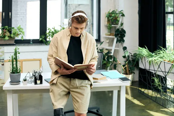 A stylish young man is reading a book while standing at his desk in a contemporary office adorned with plants. — Stock Photo