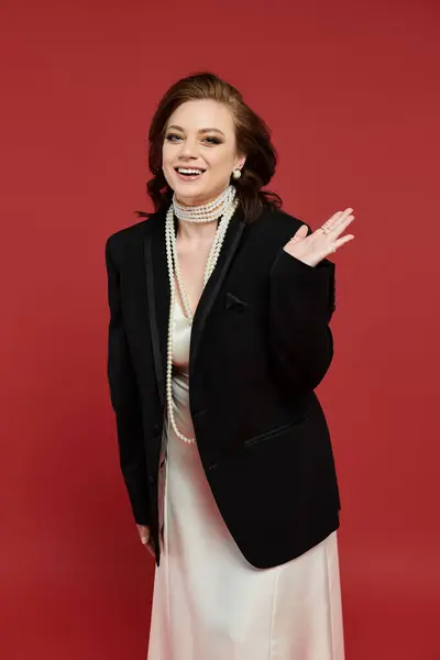 A young, beautiful woman dressed elegantly waves with a bright smile against a bold red background. — Stock Photo