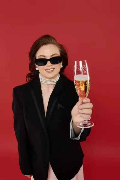 A young woman in elegant attire raises a glass, celebrating in front of a striking red backdrop. — Stock Photo
