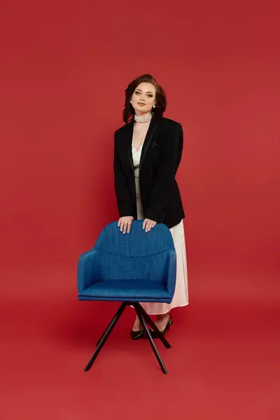 A young woman in elegant attire stands by a blue chair against a red background. — Stock Photo