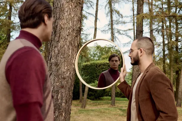 Dos hombres apuestos disfrutan del aire fresco del otoño mientras admiran sus reflejos en un espejo circular. - foto de stock