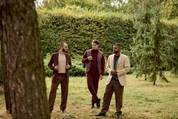 Three fashionable men confidently pose in a scenic autumn field surrounded by greenery and colorful foliage. — Stock Photo
