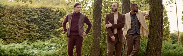 Vestido con un elegante atuendo de otoño, tres hombres están de pie con confianza en un colorido campo rodeado de naturaleza. - foto de stock