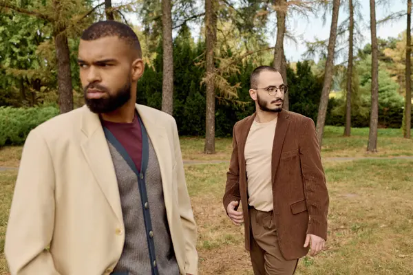 Two fashionable men enjoy a leisurely walk in an autumn field amidst colorful foliage and tall trees. — Stock Photo