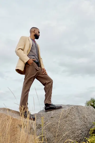 A dapper gentleman stands atop a rock, showcasing autumn fashion while surrounded by a tranquil field. — Stock Photo