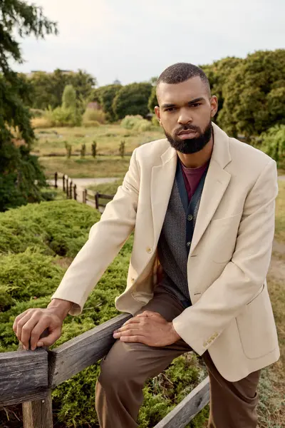 Ein fescher Mann steht selbstbewusst auf einem Holzzaun und präsentiert Herbstmode in einer ruhigen Naturkulisse. — Stockfoto