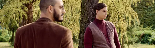 Dos hombres guapos vestidos con elegantes trajes de otoño posan elegantemente contra un telón de fondo de follaje colorido. - foto de stock