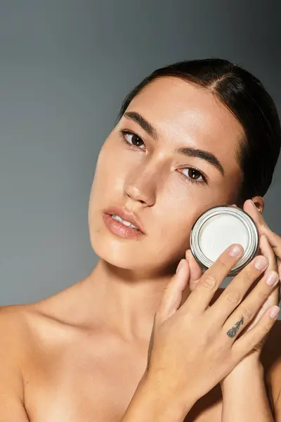 The beautiful woman gently showcases a skincare jar while highlighting her flawless complexion. — Stock Photo