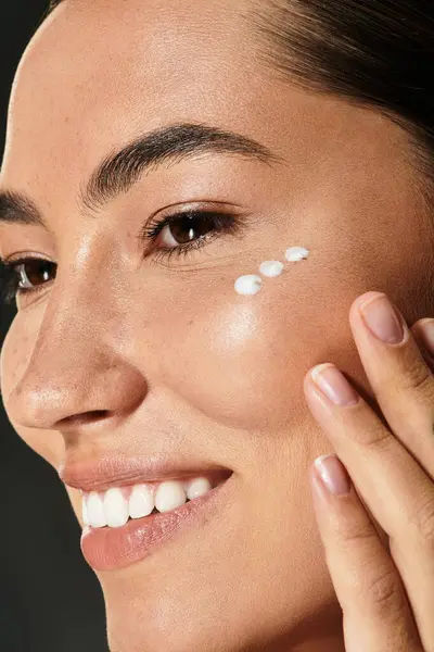 Une femme à la peau radieuse applique gracieusement de la crème sur sa joue tout en souriant. — Photo de stock