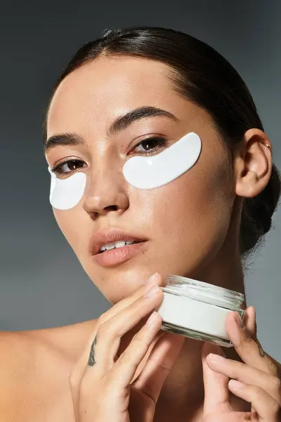 The woman elegantly holds a jar of cream while relaxing with under eye patches. — Stock Photo