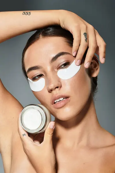 A beautiful woman applies eye patches while holding a skincare jar, exuding confidence and grace. — Stock Photo