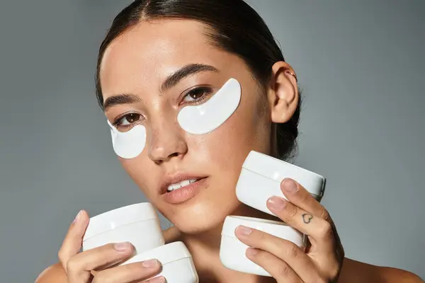 A beautiful woman applies eye masks while holding various skincare products in her hands. — Stock Photo