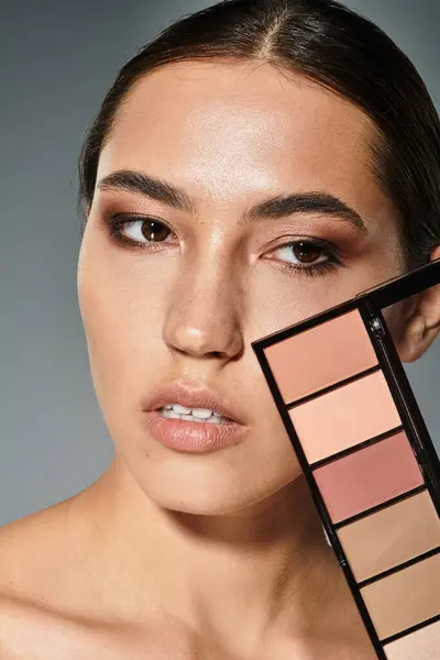 Une belle femme met en valeur ses traits tout en tenant une palette de maquillage dans un environnement élégant. — Photo de stock