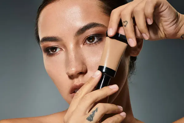 A stunning woman expertly applies foundation to enhance her natural beauty against a soft backdrop. — Stock Photo