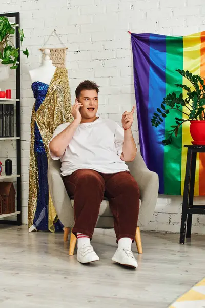 An appealing queer person sits comfortably in a stylish chair, animatedly talking on the phone. — Stock Photo