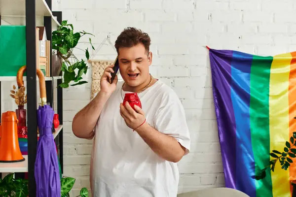 A queer individual talks on the phone, holding a red item, surrounded by plants and rainbow decor. — Stock Photo