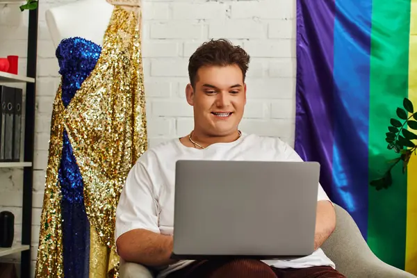 A queer person happily engages with a laptop amid vibrant decor and shining outfits. — Stock Photo