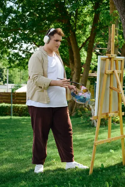 A creative person paints joyfully under trees, wearing headphones and a bright smile. — Stock Photo
