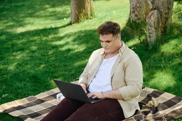 A queer person sits on a blanket, focused on their laptop in a lush green park. — Stock Photo
