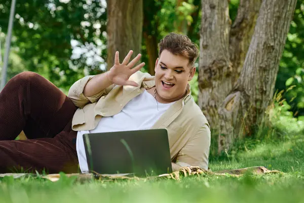 A person lounges on the grass, waving happily while interacting with a laptop outdoors. — Stock Photo