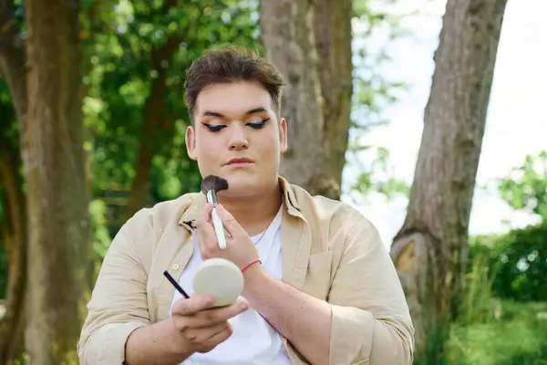 A queer person focuses on makeup application while surrounded by lush greenery and trees. — Stock Photo