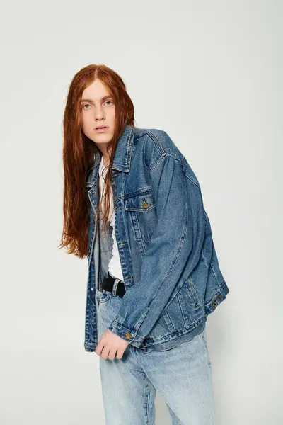 Un garçon aux longs cheveux roux saisissants présente une tenue en denim tout en posant en toute confiance dans un studio créatif. — Photo de stock