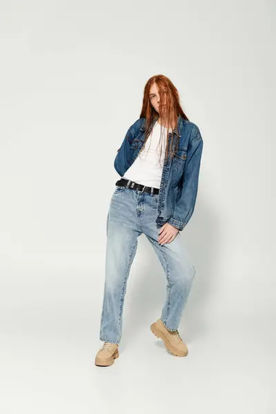 Un adolescent aux longs cheveux roux pose en toute confiance dans une tenue en denim élégant, exsudant la jeunesse. — Photo de stock