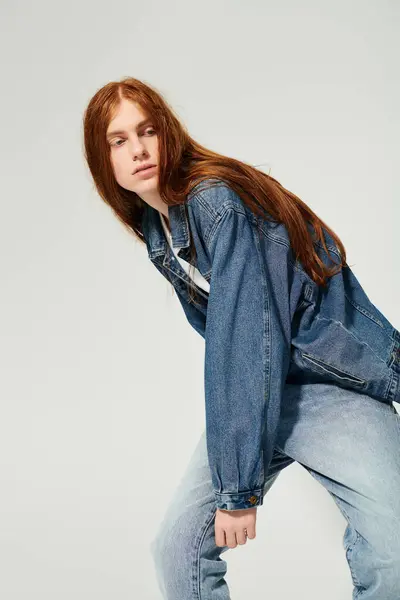 A teenage boy with striking long red hair confidently poses in a stylish denim outfit against a plain backdrop. — Stock Photo