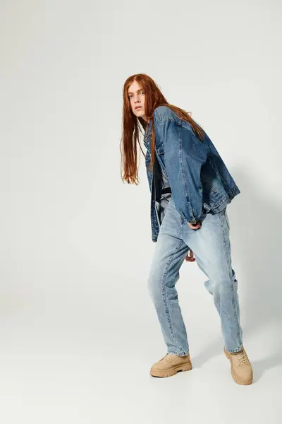 A teenage boy with striking long red hair stands poised in a denim ensemble, showcasing his unique style. — Stock Photo