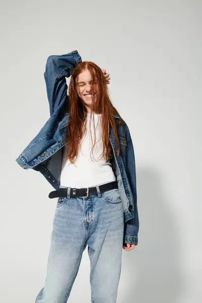 A teenage boy with flowing red hair strikes a playful pose in a trendy denim ensemble against a neutral backdrop. — Stock Photo