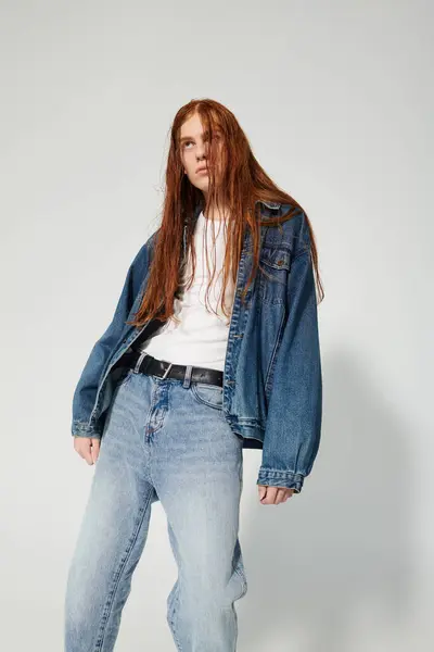 A teenage boy with striking long red hair confidently poses in a denim outfit against a studio backdrop. — Stock Photo