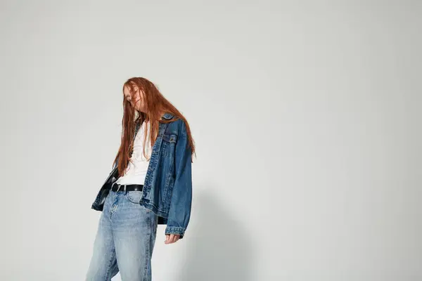 A teenage boy with flowing red hair confidently poses in a stylish denim outfit against a simple backdrop. — Stock Photo