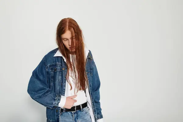 A teenage boy with long red hair confidently poses in a stylish denim outfit against a simple backdrop. — Stock Photo