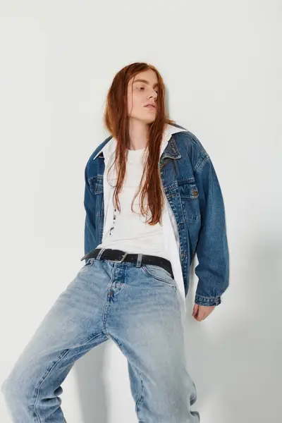 A teenage boy with long red hair confidently poses in a stylish denim outfit against a bright background. — Stock Photo