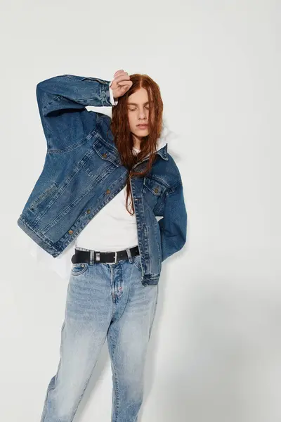 A teenage boy with long red hair strikes a confident pose in a trendy denim outfit within a minimalist studio. — Stock Photo