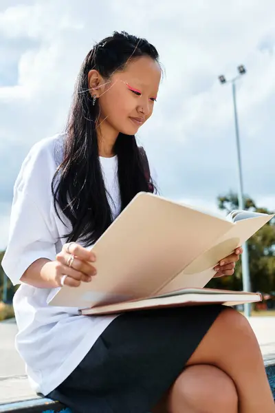 A fashionable young Asian student rests outside, engaging with her study materials under a bright sky. — Stock Photo