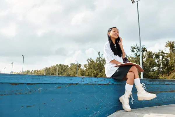 A young Asian student relaxes on a blue railing, chatting on her phone while showcasing a unique fashion sense. — Stock Photo