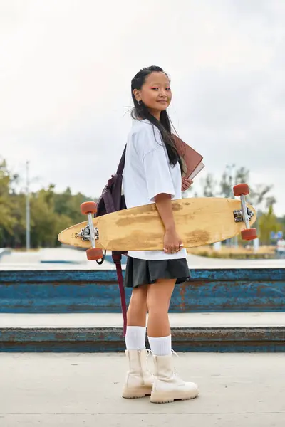 Une jeune fille asiatique en tenue tendance tient un skateboard, mettant en valeur son esprit vibrant au parc. — Photo de stock