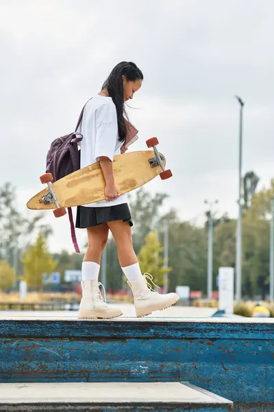 A stylish young Asian girl holds a skateboard while exploring the vibrant outdoor atmosphere of the city. — Stock Photo