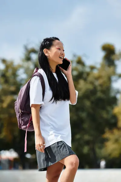 Eine junge asiatische Studentin in modischem Outfit telefoniert fröhlich, während sie durch einen sonnigen Außenbereich läuft. — Stockfoto