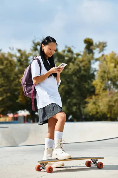 A young student girl dressed in a stylish outfit stands on her skateboard, focused on texting her friends. — Stock Photo