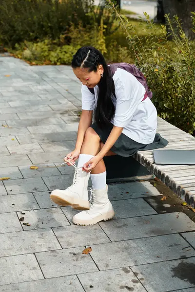 A young Asian woman sits outdoors, adjusting her stylish boots while enjoying the vibrant atmosphere of the park. — Stock Photo