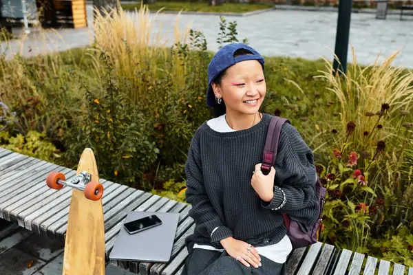A cheerful young Asian student sits outside, wearing a trendy outfit, with a skateboard beside her, enjoying the day. — Stock Photo