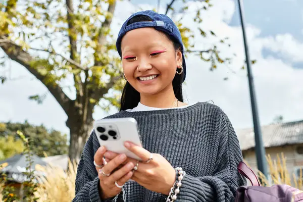 Стильная азиатская студентка улыбается во время занятий с телефоном, окруженная зеленью и яркой культурой. — стоковое фото