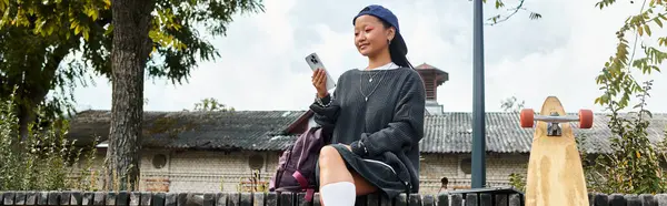 A stylish young Asian student sits casually outdoors, focused on her phone while enjoying the warm day. — Stock Photo