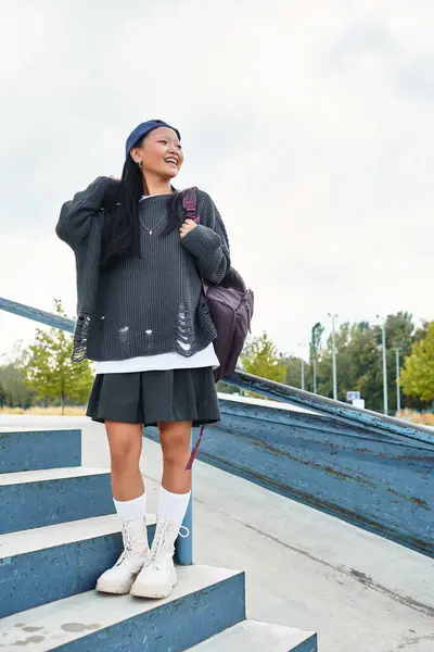 The stylish young girl with a backpack stands on the steps, smiling brightly and embracing the sunny atmosphere. — Stock Photo