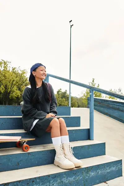 A stylish young Asian student smiles while sitting on blue steps, soaking in the sunny day outdoors. — Stock Photo