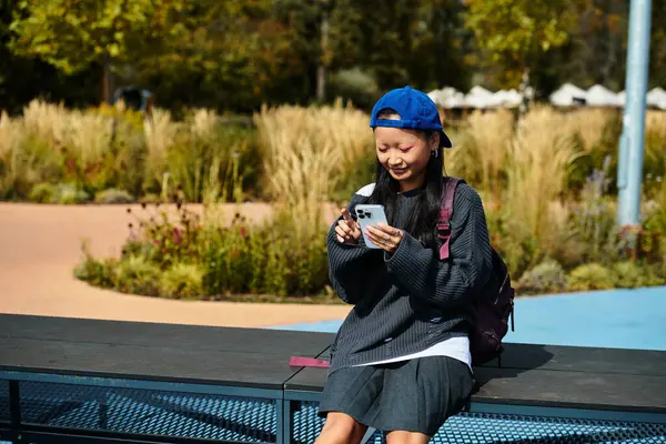A cheerful young Asian girl sits comfortably outdoors, engaged with her smartphone in a lively park setting. — Stock Photo