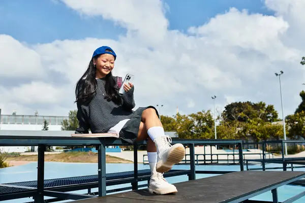 A fashionable young student girl relaxes outdoors, smiling as she uses her phone on a warm day. — Stock Photo