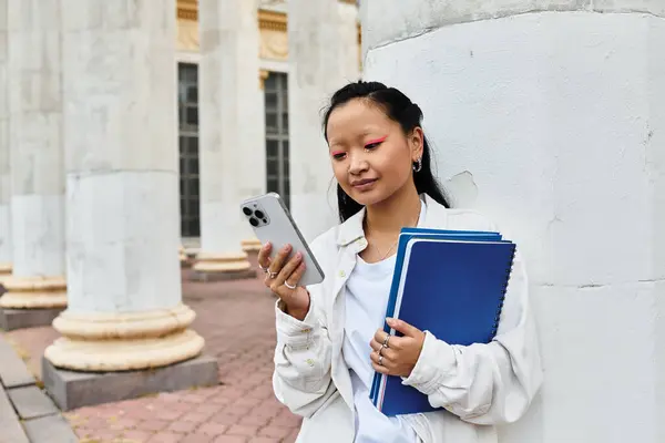 A pretty young Asian student in a fashionable outfit holds books and looks at her phone near campus pillars. — Stock Photo
